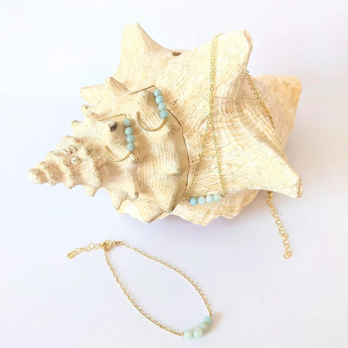 The Jordan Amazonite Collection in 14k gold filled featuring J hoop earrings, necklace and bracelet, each with four small round amazonite gemstones displayed on and around a conch shell.