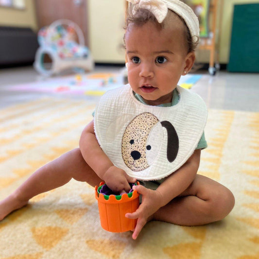 A baby wearing an ivory double cotton gauze bib with a puppy applique is sitting on the floor, holding a small orange toy bucket.