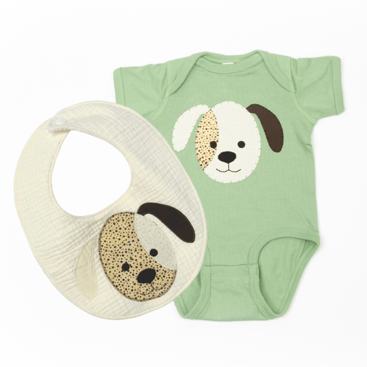 A baby wearing an ivory double cotton gauze bib with a puppy applique is sitting on the floor, holding a small orange toy bucket.