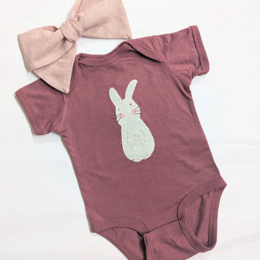 A baby sitting on the floor wearing apink gingham tieable headband and a rouge onesie with a hand-appliquéd white and pink bunny