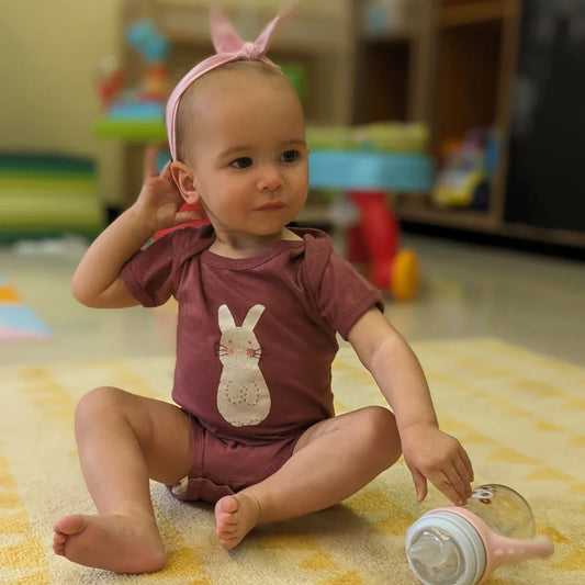 A baby sitting on the floor wearing apink gingham tieable headband and a rouge onesie with a hand-appliquéd white and pink bunny