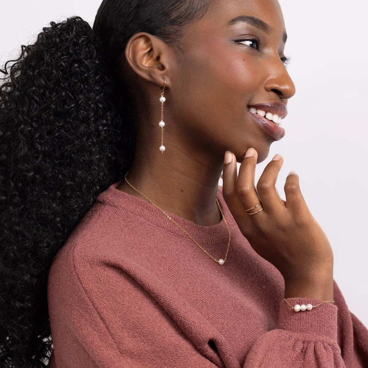 Woman wearing a pink sweater wearing freshwater pearl triple drop leverback earrings, necklace, and bracelet in 14k gold filled on a white background