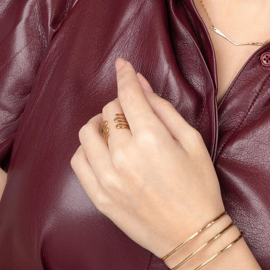 A woman's hand wearing a brass, adjustable Journey of Hope ring, with part of a leather jacket and gold-colored jewelry visible in the background.
