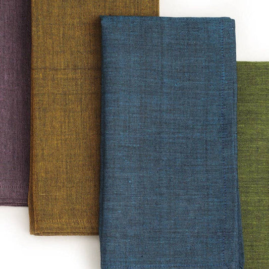 Four folded textured fabric napkins in overlapping vertical rows, shown on a plain white background. The napkins are in muted purple, mustard brown, slate blue, and olive green.