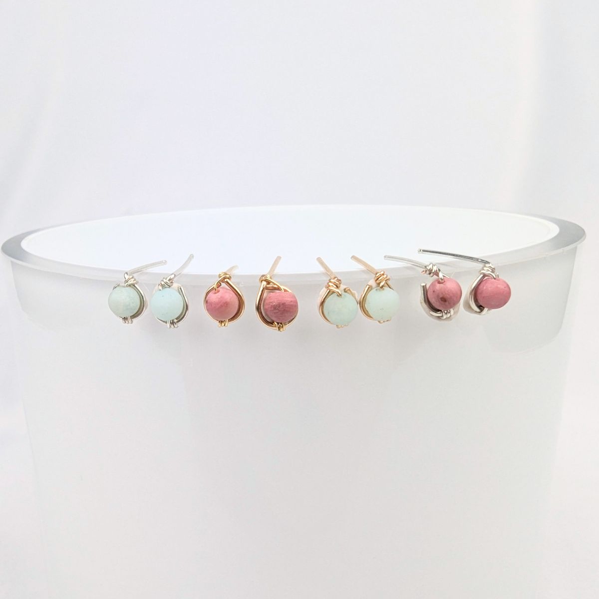 A set of small teardrop-shaped drop earrings displayed across the rim of a white cylindrical stand. The earrings feature pastel mint green and dusty rose beads with gold-toned settings.