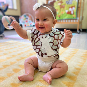 A baby sitting on the floor wearing a giraffe print bib with off-white giraffe applique and a plush terry cloth back.