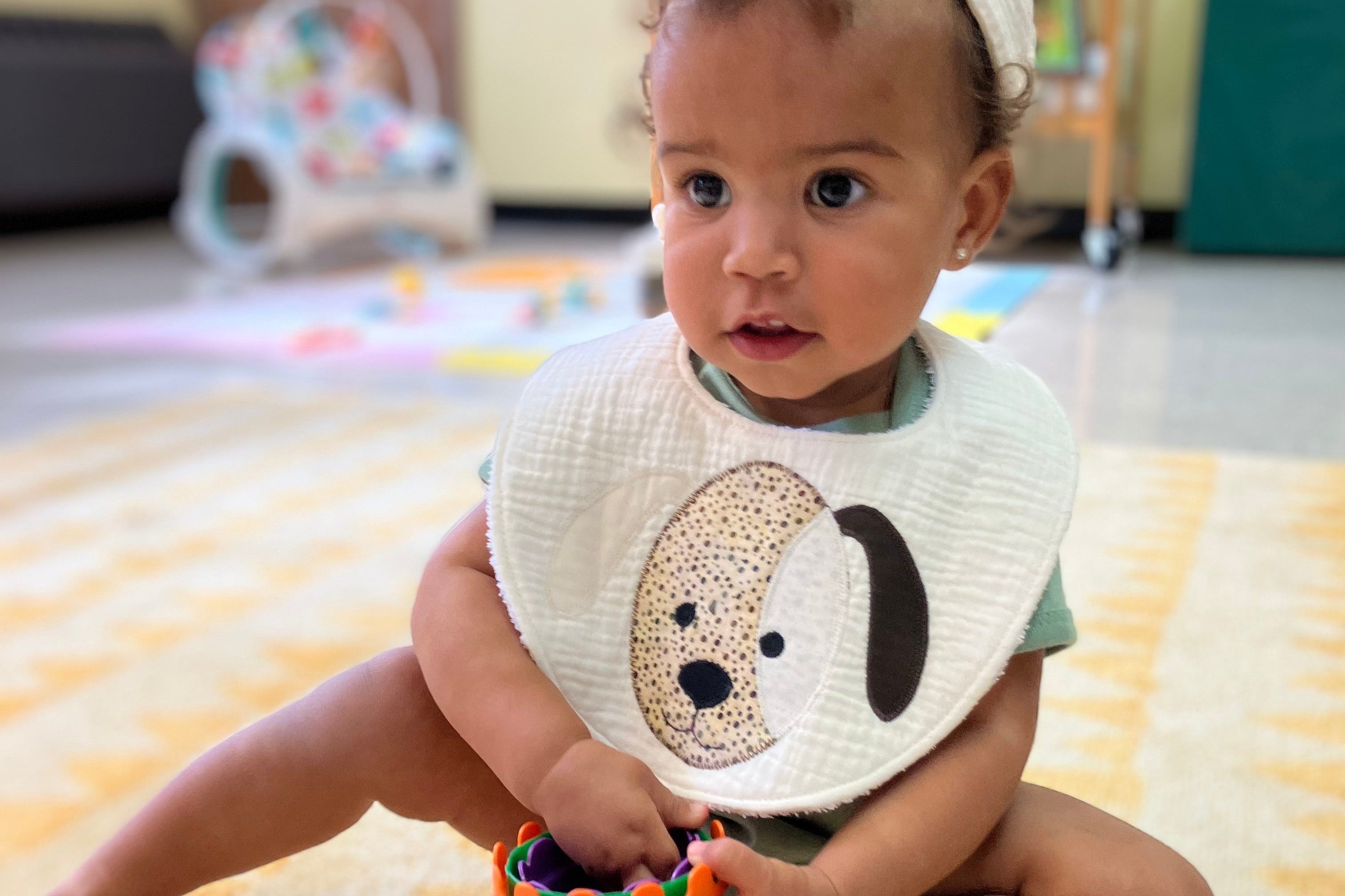 Baby sitting on a rug wearing a bib with a dog design, holding an orange toy.