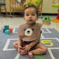 Baby sitting on a colorful play mat wearing a bear-themed bib in a nursery setting.