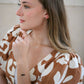 A woman in a brown and white patterned top wearing delicate gold jewelry, including a thin gold chain bracelet with small black onyx beads, a gold ring, and a gold necklace, shown in a close-up profile shot.
