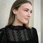 A woman with light brown hair wearing a black lace mock-neck top and a small black stud earring, shown in a close-up profile against a light gray stone wall.