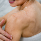 Woman smiling while applying Thistle Farms exfoliating body scrub to her upper arm and shoulder.
