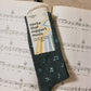 Dark gray sock with musical note design on a sheet of music, featuring a 'conscious step' tag.