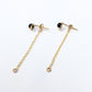 A pair of gold dangle earrings with small black bead studs and delicate chain drops, displayed on a white textured fabric background.
