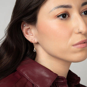 A woman wearing the Mini-hoop Aqua Quartz Charm Earrings in 14k Gold Filled, stacked with an ear cuff and huggie earring.