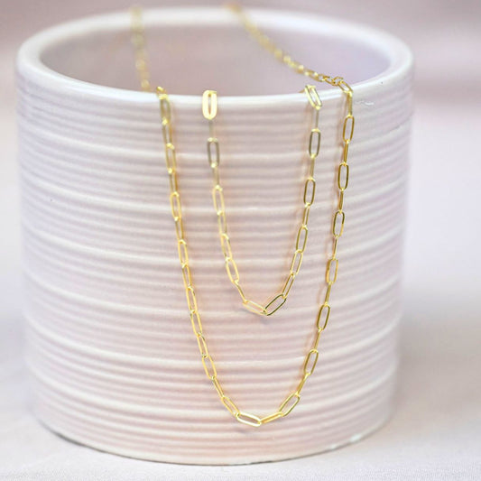a gold paperclip chain necklace and bracelet draped over the edge of a pink ceramic cup on a pink backdrop