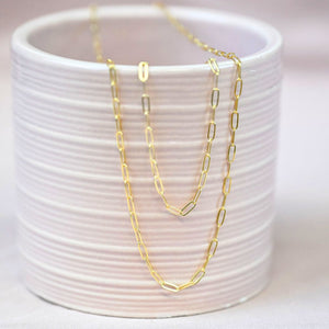 a gold paperclip chain necklace and bracelet draped over the edge of a pink ceramic cup on a pink backdrop