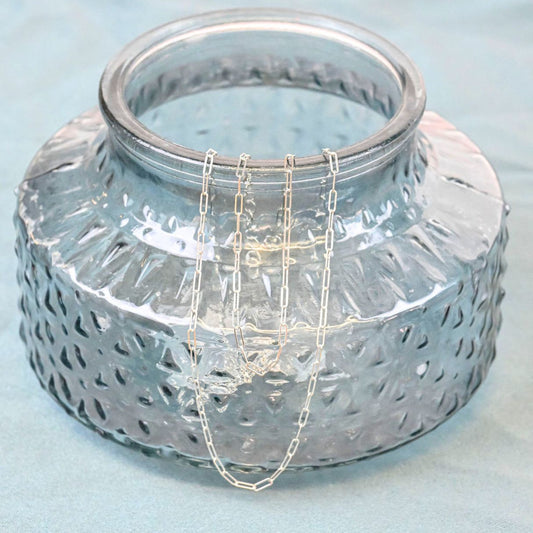 a sterling silver paperclip chain necklace and bracelet draped over the opening of a blue glass vase on a blue background