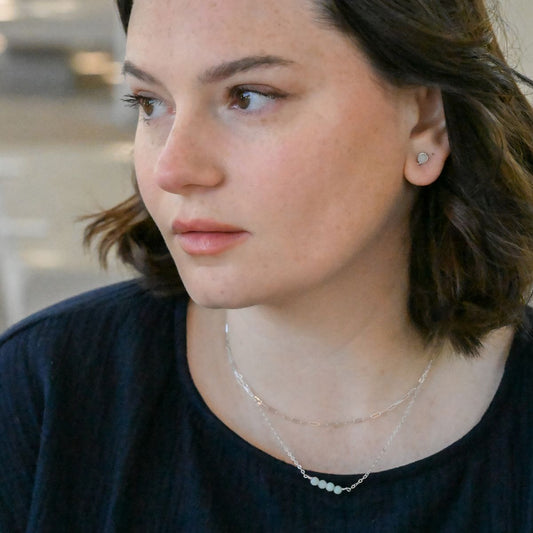 brunette model wearing silver stud earrings with a blue green bead, as well as two silver necklaces