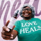 Person holding a bottle of Thistle Farms hand sanitizer with 'Love Heals' shirt in the background