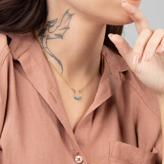 A close-up of a woman wearing a beige shirt and the Moab Western Beaded Necklace in 14k gold filled. Horseshoe design with turquoise beads.