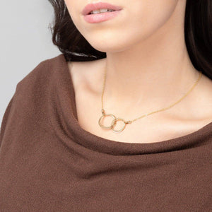 The Zomi Circles of Unity Necklace in 14k gold filled features interlocking circles and is modeled by a young woman in a brown sweater.