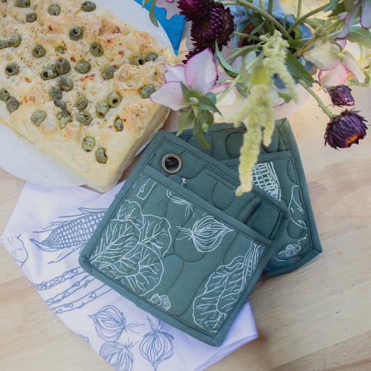 Two dark green quilted pot holders with white line-art vegetable prints, including leafy greens, onions, and corn, displayed on a white background.