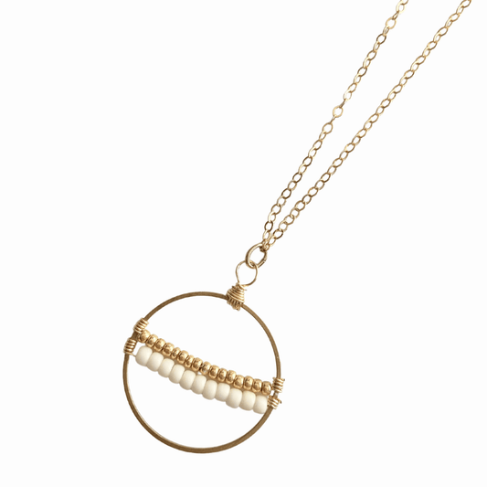 A woman wearing the Samira Seeds of Hope Necklace in 14k Gold Filled which features a brass circle with white and gold seed beads threaded across the center.