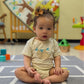 Child wearing beige onesie with sloth design sitting on a colorful mat in a playroom with toys around