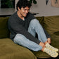 Man sitting on a green couch wearing a gray sweater and light blue jeans, smiling, while putting on yellow ankle socks with smiley faces.