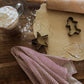 A styled flat-lay of baking tools on a wooden table, featuring metal star and gingerbread cookie cutters on a piece of rolled-out dough, a wooden rolling pin, a glass mug filled with whipped cream, and a red-and-white striped cloth.