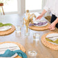 Person setting a table with place settings and glasses in a bright room.