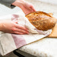 A freshly baked round loaf of rustic bread resting on a wooden cutting board, wrapped in a cream and mauve striped kitchen towel, with a person's hands holding it on a speckled white countertop.