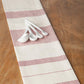 A striped kitchen towel or napkin in cream and dusty pink, laid diagonally on a wooden table with a small set of white measuring spoons resting on top.